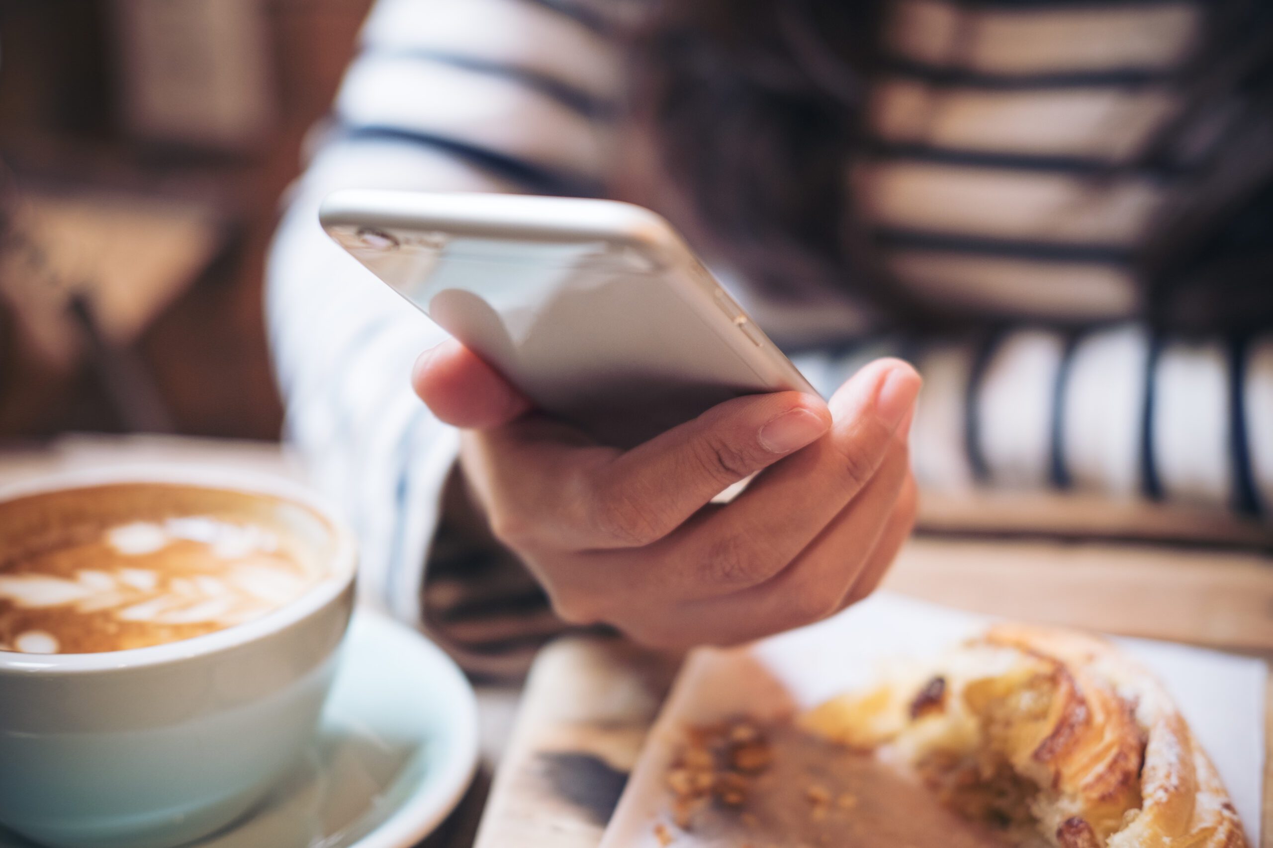 Closeup image of a woman's hands holding and using smart phone with latte coffee cup and bread on wooden table in vintage cafe