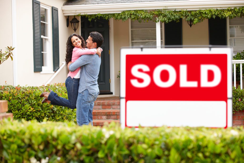 A couple hugging outside of their home with a sold sign in front of them.  Loan for homebuyers